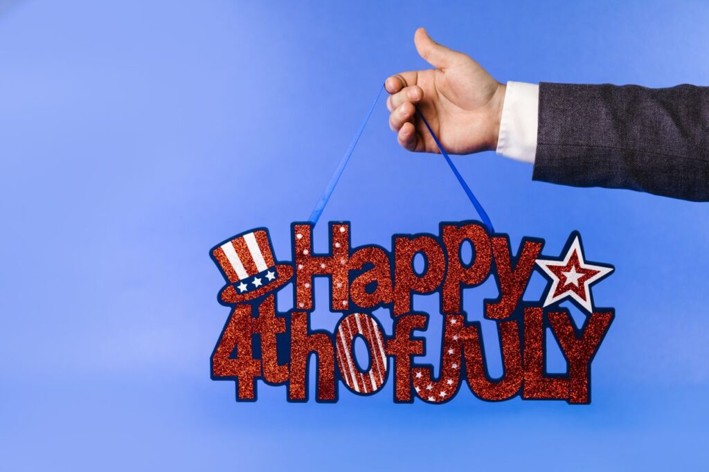 Hand holding glittery 4th of July sign with stars and stripes on blue backdrop.